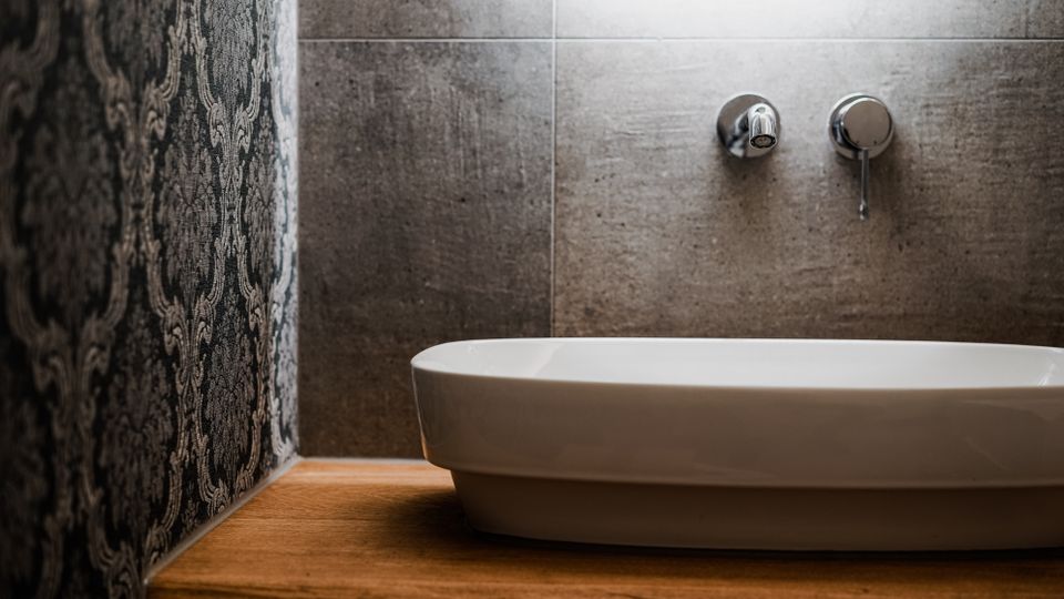 Modernes Badezimmer-Waschbecken aus weißer Keramik auf einer Holzablage, davor eine Wand aus grauen Fliesen mit wandmontiertem Wasserhahn und Bedienhebel. Links ist eine dunkle, gemusterte Tapete als Kontrast zur Fliesenwand zu sehen.