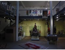 Innenraum eines Museums mit Puppen und Marionetten. Mehrere Glasvitrinen zeigen Figuren, auf zwei Etagen mit Metallgeländern verteilt; im Vordergrund liegt ein Teppich, und die Beleuchtung stammt von Deckenstrahlern.