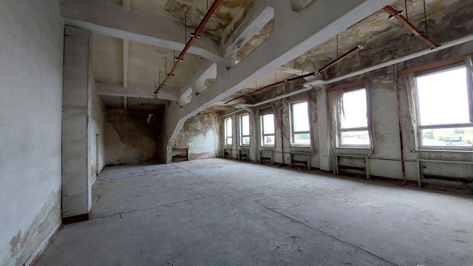 Ein leerer, verfallener Raum mit Betonboden und abgenutzten Wänden. Die Decke zeigt freiliegende Rohre, und an einer Seite befinden sich mehrere große Fenster mit Blick auf eine Außenlandschaft.