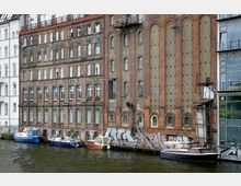 Backsteinfassade eines historischen Gebäudes am Wasser, teilweise mit Graffiti bedeckt, daneben moderne und ältere Architektur. Vor dem Gebäude liegen mehrere Boote am Ufer.