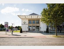 Moderne, weiß gestrichene Gebäude mit dem Schriftzug "Klubhaus" an der Fassade, großen Fenstern und einem breiten Eingang; davor ein gepflasterter Vorplatz mit Bäumen, Wegweisern und einer Parkuhr. Im Hintergrund sind Wohnhäuser und Grünflächen sichtbar.