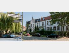 Straßenszene mit einem historischen Gebäude im Hintergrund, das weiße Fassaden mit rotbraunen Fensterrahmen und Verzierungen hat, sowie mehreren Fenstern und einem kleinen Turm. Im Vordergrund sind eine Kopfsteinpflasterstraße, parkende Autos und Straßenschilder mit den Namen „Freiwaldauer Weg“ und „Geibelstraße“.