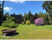 Großer Garten mit weitläufiger Rasenfläche, umgeben von hohen Bäumen und Sträuchern; im Vordergrund steht eine runde Feuerschale auf einem Steinpodest. Im Hintergrund ein kleines Haus mit Reetdach sowie blühende Büsche, darunter ein großer rosa Rhododendron.