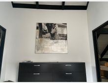 Ein modernes Zimmer mit weißen Wänden und einer schwarzen Kommode, auf der ein paar kleine Gegenstände liegen. Darüber hängt ein Schwarz-Weiß-Kunstbild mit einer Person und einem Schriftzug an der Wand.