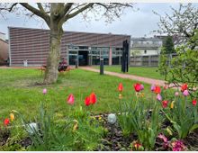 Ein moderner, einstöckiger Gebäudekomplex mit einer Fassade aus horizontalen Metallstreifen, umgeben von einer grünen Rasenfläche mit bunten Tulpen im Vordergrund. Ein einzelner Baum und ein gepflasterter Weg ergänzen die Parklandschaft, während im Hintergrund Häuser und Zäune sichtbar sind.
