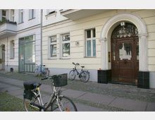 Mehrstöckiges Wohngebäude mit heller Fassade, verzierten Fenstern und einem Holzeingangstürbogen mit Säulen. Vor dem Gebäude stehen mehrere Fahrräder auf einem gepflasterten Gehweg.