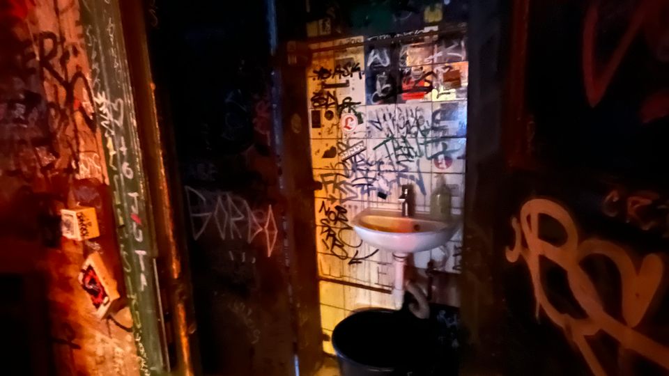 Schmaler, schwach beleuchteter Toilettenraum mit weißen Bodenfliesen und einem kleinen Waschbecken an einer gefliesten Wand. Wände und Türrahmen sind dicht mit Graffiti und Aufklebern bedeckt, daneben steht ein schwarzer Mülleimer.