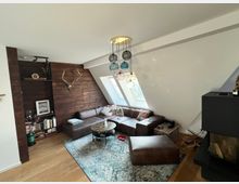 Dachgeschoss-Wohnzimmer mit schrägen weißen Wänden, einer braunen Ledercouch, einem Holzboden und einem kleinen Couchtisch. An der linken Wand befindet sich eine Holzverkleidung mit einem Regal, das Bücher und Dekor enthält, sowie ein Geweih an der Wand. Rechts steht ein moderner schwarzer Kamin, und eine Lampe mit runden Glasanhängern hängt von der Decke.