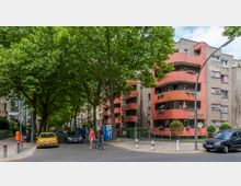 Wohnstraße mit Bäumen, die die Straße säumen, und einem mehrstöckigen Wohngebäude mit halbkreisförmigen orangenen Balkonen. Auf der rechten Seite sind geparkte Autos und ein Straßenschild, links eine gelbe Telefonzelle und Fußgänger auf dem Gehweg zu sehen.