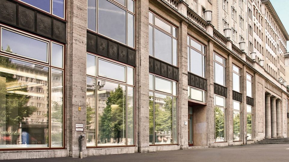 Facade of a large stone building with tall rectangular panes of reflective glass windows and decorative metal panels above them. The structure features classical architectural elements, such as columns and a balustrade, and is situated along a paved walkway.