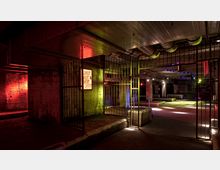 Untergrundraum mit industriellem Design, sichtbaren Rohren und Betonwänden, beleuchtet durch rote, gelbe und blaue Akzentbeleuchtung. Metallgitter und vereinzelte Betonblöcke bilden den offenen Bereich, der durch das farbige Licht betont wird.