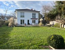 Zweistöckiges weißes Wohnhaus mit roten Dachziegeln, großen Fenstern und einer angrenzenden Terrasse. Im Vordergrund liegt ein großer, gepflegter Garten mit Büschen, Bäumen und Gräsern.