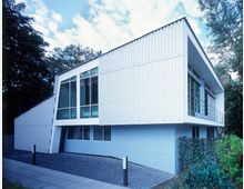 Modernes Gebäude mit einer weißen Metallfassade und großen, teilweise bodentiefen Fenstern. Umgebung aus Gehwegplatten, Kiesflächen und dichter grüner Vegetation, mit Bäumen im Hintergrund und blauem Himmel darüber.