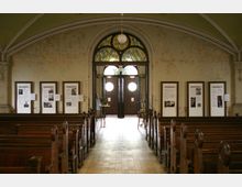 Innenraum einer historischen Kapelle mit hölzernen Kirchenbänken und schlichter, abgenutzter Wandgestaltung. Im Hintergrund befinden sich doppelflügelige Türen mit runden Fenstern und darüber ein buntes Bleiglasfenster; an den Wänden hängen gerahmte Informationstafeln.