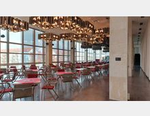 Moderner Speisesaal mit großen Glasfenstern, durch die Tageslicht einfällt. Der Raum verfügt über Tische mit roten Tischläufern, dazu passende Stühle mit roten Polstern, hängende Metallleuchten an der Decke und helle Steinwände.