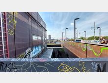 Blick auf einen S-Bahnhof mit Gleisen unter einer Brücke, flankiert von modernen braunen Fassaden mit Graffiti. Im Hintergrund sind Stadtgebäude, ein hoher Turm und Oberleitungsmaste zu sehen, während Passanten auf der Brücke entlanggehen.