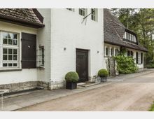 Weiß verputztes Landhaus mit dunklem Holzdach und Fachwerkakzenten, flankiert von großen Holzfenstern mit Fensterläden und dekorativen Pflanzenkübeln. Der Eingangsbereich zeigt eine dunkle Holztür mit Metallverzierungen, umgeben von gepflastertem Weg und begrünten Elementen.