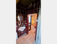 Ein kleines Badezimmer mit dunkelroten Fliesen und weißem Fugenmuster; links ein Wandspiegel mit vergoldetem Rahmen und ein kleines Waschbecken mit dekorativen Halterungen. Durch eine offene Tür sieht man einen angrenzenden Raum mit Teppichboden, einem orangefarbenen Bett und Vorhängen.