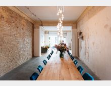 Langer Raum mit unverputzter Backsteinwand und heller Putzwand, einem massiven Holztisch mit blauen Stühlen und einer Hängelampe aus Glassphären. Im Hintergrund ein heller Wohnbereich mit Pflanzen, Sofas und großen Fenstern.