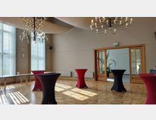 Ein heller Veranstaltungsraum mit Parkettboden, hohen Fenstern mit vertikalen Lamellen und zwei Kronleuchtern an der Decke. Im Raum stehen hohe, mit Stoff bedeckte Stehtische in Rot und Schwarz, und im Hintergrund sind Holztüren mit Glaseinsätzen zu sehen, die zu einem weiteren Bereich mit Pflanzen führen.