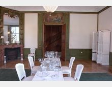 Elegantes Zimmer mit klassischer Einrichtung, bestehend aus einem Kamin mit Marmorumrandung, einer gedeckten Tafel mit weißen Stühlen und einer hölzernen Tür, die zu einer Treppe führt. Die Wände sind teilweise grün-weiß mit goldenen Verzierungen und einer Deckenkunstszene oberhalb der Tür.