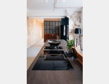 Moderne Küche mit einer langen Arbeitsplatte aus dunklem Material, einem eingebauten Kochfeld und einem Wasserhahn. Im Hintergrund sind freiliegende Ziegelwände, eine Holztür mit Glasfenstern und eine Wand mit teilweise abblätterndem Putz sichtbar.