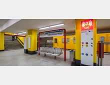 U-Bahnhof Jakob-Kaiser-Platz in Berlin mit gelben Wänden, modernem SOS-Notrufterminal und Sitzbänken. Im Hintergrund fährt eine gelbe U-Bahn der Linie U7 ein; rechts führen Treppen zu einem Ausgang.