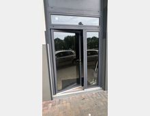 Eine moderne Glastür mit einem grauen Rahmen, teilweise geöffnet, eingebettet in eine graue Gebäudefassade. Im Hintergrund sind Bäume, parkende Autos und ein gepflasterter Boden sichtbar.