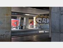 Bahnhofsunterführung mit Betonstützen und Graffiti an den Wänden, im Hintergrund ein Bahnsteig mit einer vorbeifahrenden roten Regionalbahn. Schilder, ein Abfallbehälter und ein abgestelltes Fahrrad ergänzen das Stadtbild.