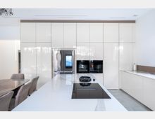 Moderne Küche mit glänzenden weißen Einbauschränken, einem großen Kühlschrank mit integriertem Bildschirm, schwarzen Einbaugeräten sowie einer Kücheninsel mit weißer Steinplatte und schwarzem Kochfeld. Im Vordergrund befindet sich ein Esstisch mit gepolsterten Stühlen.