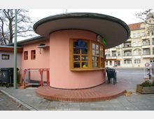 Kleines rundes Gebäude mit rosa Fassade, großen Holzfenstern und einem flachen, weit überhängenden Dach. Die Umgebung zeigt eine Stadtstraße mit Bäumen und mehrstöckigen Wohnhäusern im Hintergrund.