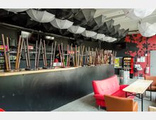 Barbereich mit hochgestellten Stühlen auf einer langen Theke, darüber eine Decke mit schwarzen und weißen dekorativen Regenschirmen. Im Vordergrund befinden sich eine rote Couch, ein Holztisch und braune Stühle; im Hintergrund Automaten und eine Wand mit rotem Blättermuster.