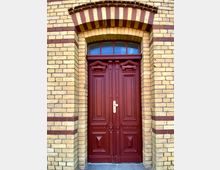 Rote Holztür mit dekorativen Paneelen und Messinggriff, eingefasst in helle gelblich-beige Backsteinmauern mit roten Zierstreifen; darüber ein halbkreisförmiges Fenster mit Sprossen.