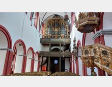 Innenansicht einer historischen Kirche mit weiß gestrichenen Wänden, roten Backsteinbögen und Holzdecke. Im Vordergrund ist eine reich verzierte, goldene Kanzel zu sehen, während im Hintergrund eine große Orgel mit vergoldeter Dekoration und darüber ein gotisches Fenster erkennbar sind.