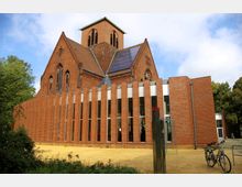 Kirchengebäude aus rotem Backstein mit einem steilen Dach und einem Turm, ergänzt durch eine moderne Fassade mit vertikalen Backsteinsäulen und großen Fenstern. Im Vordergrund befindet sich ein kleiner Platz mit Fahrradständer, umgeben von Bäumen.