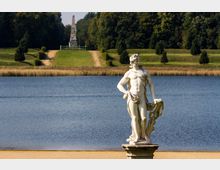 Eine weiße Marmorskulptur eines nackten Mannes steht auf einem Sockel im Vordergrund, am Rand eines ruhigen Sees. Im Hintergrund führen symmetrisch gestaltete Wege durch eine grüne Parklandschaft zu einem Obelisken.