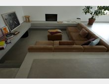 Ein minimalistisches Wohnzimmer mit einer versenkten braunen Polstercouch in U-Form, einem beigen Teppich und einem passenden Hocker in der Mitte. Im Hintergrund befinden sich ein Kamin, ein Betonregal mit Büchern und Deko sowie eine große Zimmerpflanze in einem Topf.