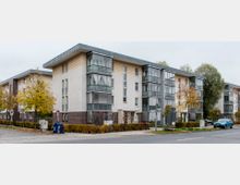 Moderne Wohngebäude mit drei Etagen, einem Mix aus verputzten und verklinkerten Fassaden sowie großen verglasten Balkonen. Die Straße ist von Herbstbäumen und geparkten Autos gesäumt, und im Vordergrund sind Straßenschilder und eine Bushaltestelle sichtbar.