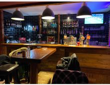 Ein kleiner, gemütlicher Barbereich mit einer Holztheke, Zapfhähnen und Flaschenregalen an einer Wand aus schwarzen, glänzenden Kacheln. Im Vordergrund steht ein Holztisch mit Stühlen, während ein Fernseher im Hintergrund ein Sportprogramm zeigt.