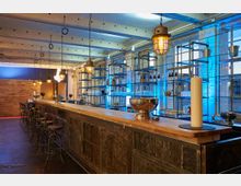 Industriell gestaltete Bar mit einer langen Holztheke und Hockern aus Metall, umgeben von Regalen mit Flaschen und Gläsern. Die Szenerie wird durch warme Pendelleuchten und blaue Hinterleuchtung der Fenster akzentuiert.