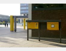 Außenbereich mit gelben Briefkästen an einer dunklen Backsteinwand; im Hintergrund eine offene Fläche mit Überdachung, gelben Säulen und einem Bürogebäude mit modernen Glasfassaden. Zwei Personen sind in der Ferne auf der linken Seite zu sehen.