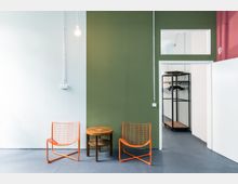 Minimalistisch eingerichteter Raum mit einer weißen und grünen Wand, zwei orangefarbenen Drahtstühlen und einem runden Holztisch in der Mitte. Im Hintergrund ein offener Durchgang zu einem Bereich mit einer Gitter-Garderobe.