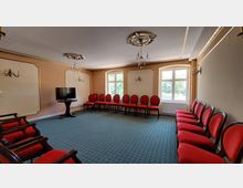 Ein eleganter Seminar- oder Besprechungsraum mit beige-goldenen Wänden und großer Fensterfront, bestuhlt mit roten Polsterstühlen in einem Halbkreis. Der Raum verfügt über verzierte Deckenleuchter, Wandlampen im klassischen Stil und einen kleinen Fernseher auf einem schwarzen Standfuß.