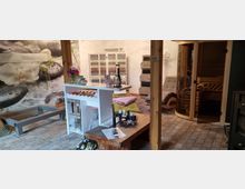 Raum mit Wellness-Ausstattung, einschließlich einer Glas-Saunakabine, einer Holzliege und Entspannungselementen wie Kissen und Dekoration. Im Vordergrund stehen ein Holz- und ein weißes Regal mit Kerzen, Ölen und Buddha-Statuen, während die Wand eine großformatige Blüten- und Stein-Grafik zeigt.