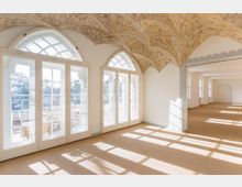 Heller Innenraum mit verzierten Gewölbedecken, großen bogenförmigen Fenstern mit Sprossen und einem Ausblick auf eine Terrasse. Der Raum ist mit einem beigefarbenen Teppichboden und minimaler Dekoration gestaltet.