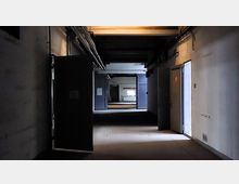 Dunkler Flur in einem Gebäude mit offenen Türen, freiliegenden Kabeln an den Wänden und einem Fenster im Hintergrund, das schwaches Licht hereinlässt. Das Umfeld wirkt industriell und verlassen.