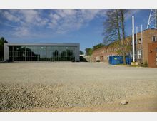 Moderne Industrieanlage mit einem großen Gebäude mit Glasfassade im Vordergrund und einem länglichen, ziegelroten Fabrikgebäude mit Schornsteinen auf der rechten Seite. Im Vordergrund befindet sich eine unbefestigte Kiesfläche, im Hintergrund sind Bäume und blauer Himmel zu sehen.