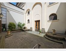 Innenhof mit gepflastertem Boden, flankiert von Pflanzen in Töpfen und erhöhten Beeten. Die helle Fassade eines Gebäudes mit Rundbogenfenstern und einer Holztür bildet den Hintergrund, teilweise in Sonnenlicht getaucht.