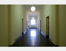 Ein langer, schlichter Flur mit gelb gestrichenen Wänden und dunklem Boden, mehreren Türen an beiden Seiten, runden Deckenleuchten und einem großen Fenster am Ende des Gangs.