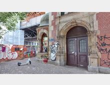 Historisches Backsteingebäude mit aufwendig verzierten Tür- und Fensterbögen, Graffiti an den Wänden und einem E-Scooter im Vordergrund. Im Hintergrund ist eine Baustelle mit Gerüsten sowie eine überdachte Metallkonstruktion sichtbar.
